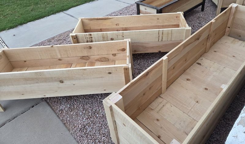 A batch of cedar planters, square and long, lined up on a gravel driveway